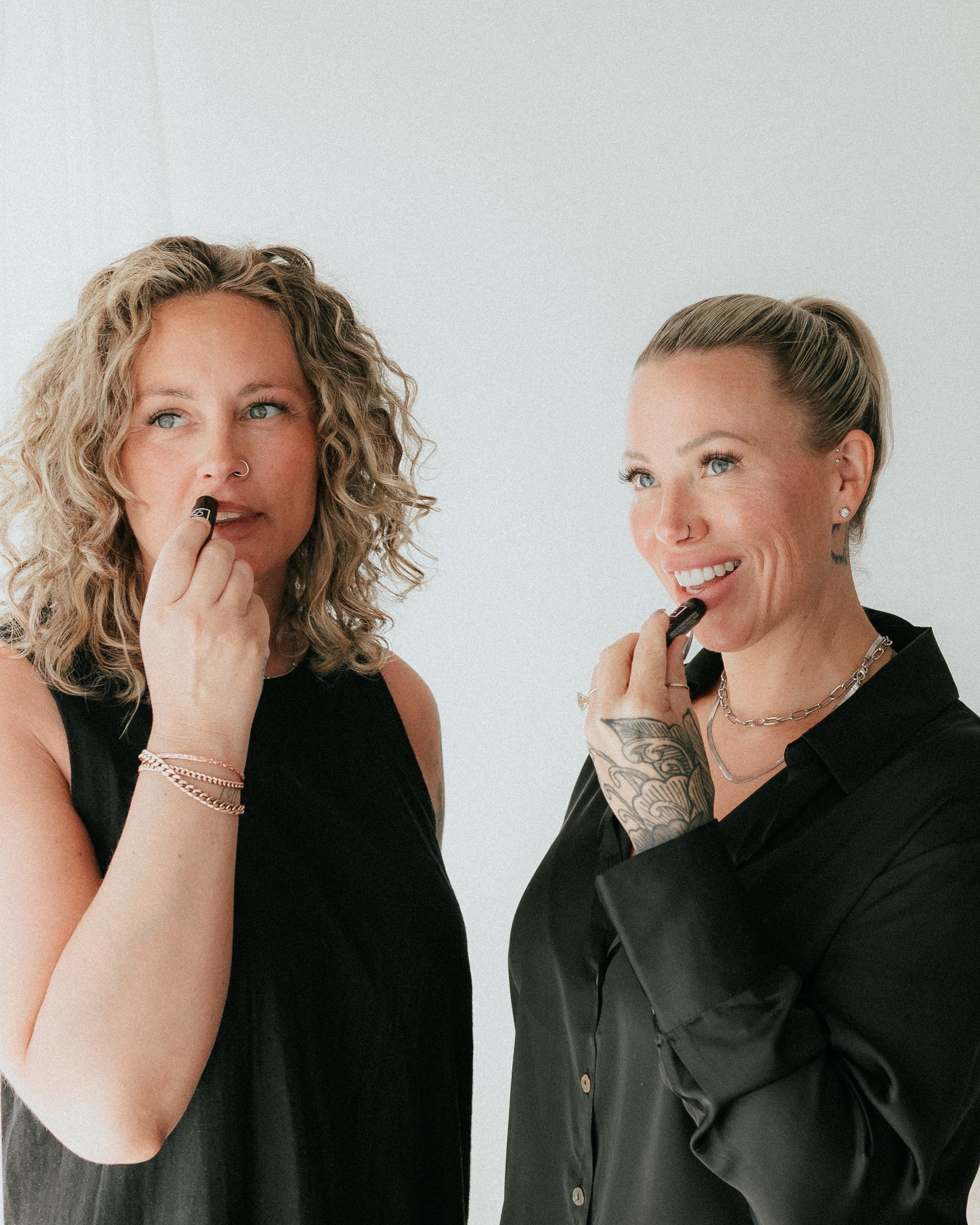 Two women standing indoors and applying lipstick. Both are wearing black tops, with one in a sleeveless black dress and the other in a black button-up shirt.