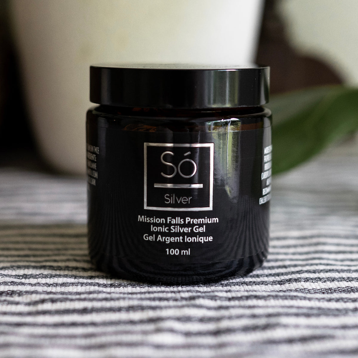 A 100 ml black jar of Só Silver Mission Falls Premium Ionic Silver Gel, placed on a gray and white striped fabric surface with a blurred background.