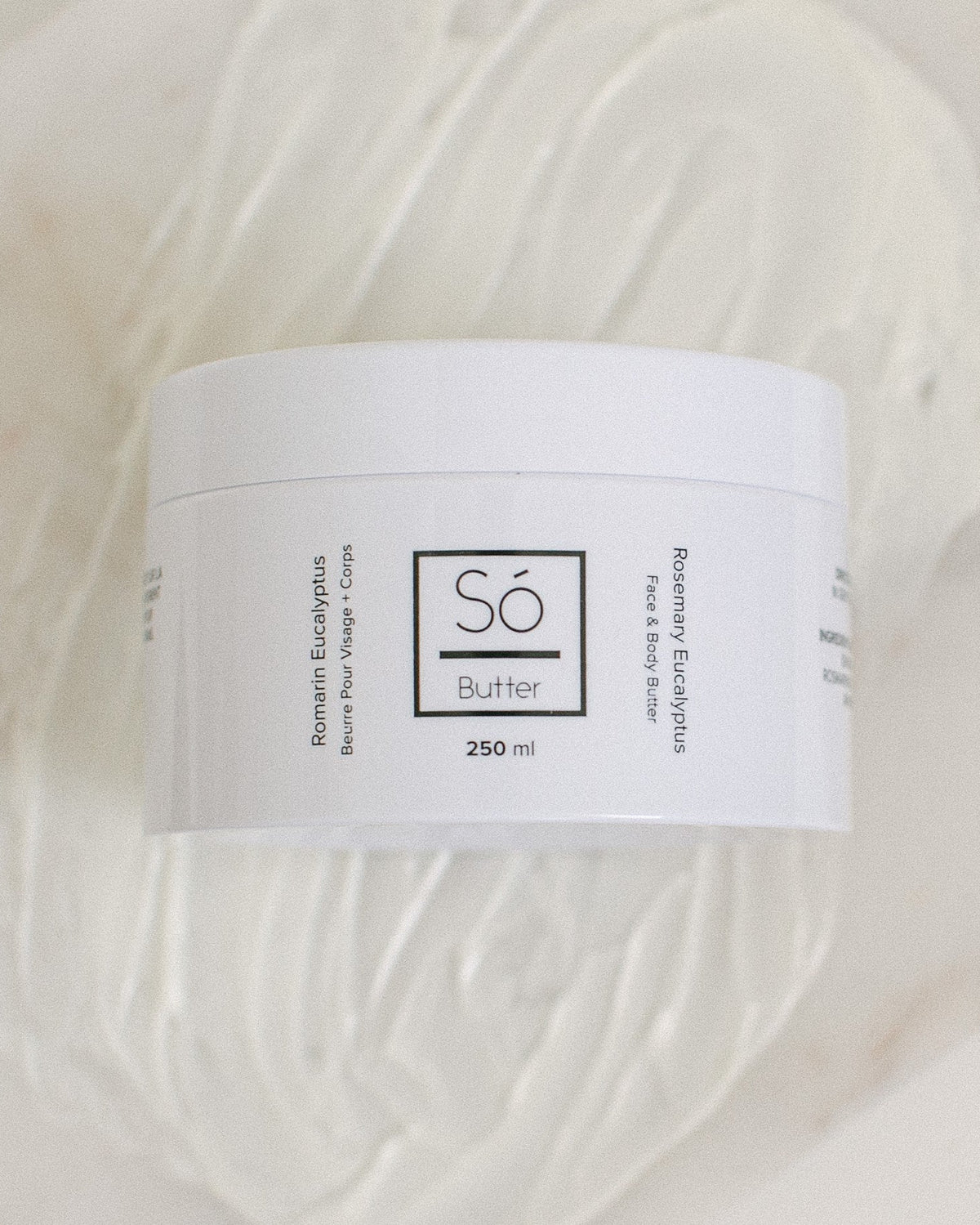A white plastic jar of Só Butter Rosemary Eucalyptus body butter, placed on top of a textured background of white whipped product