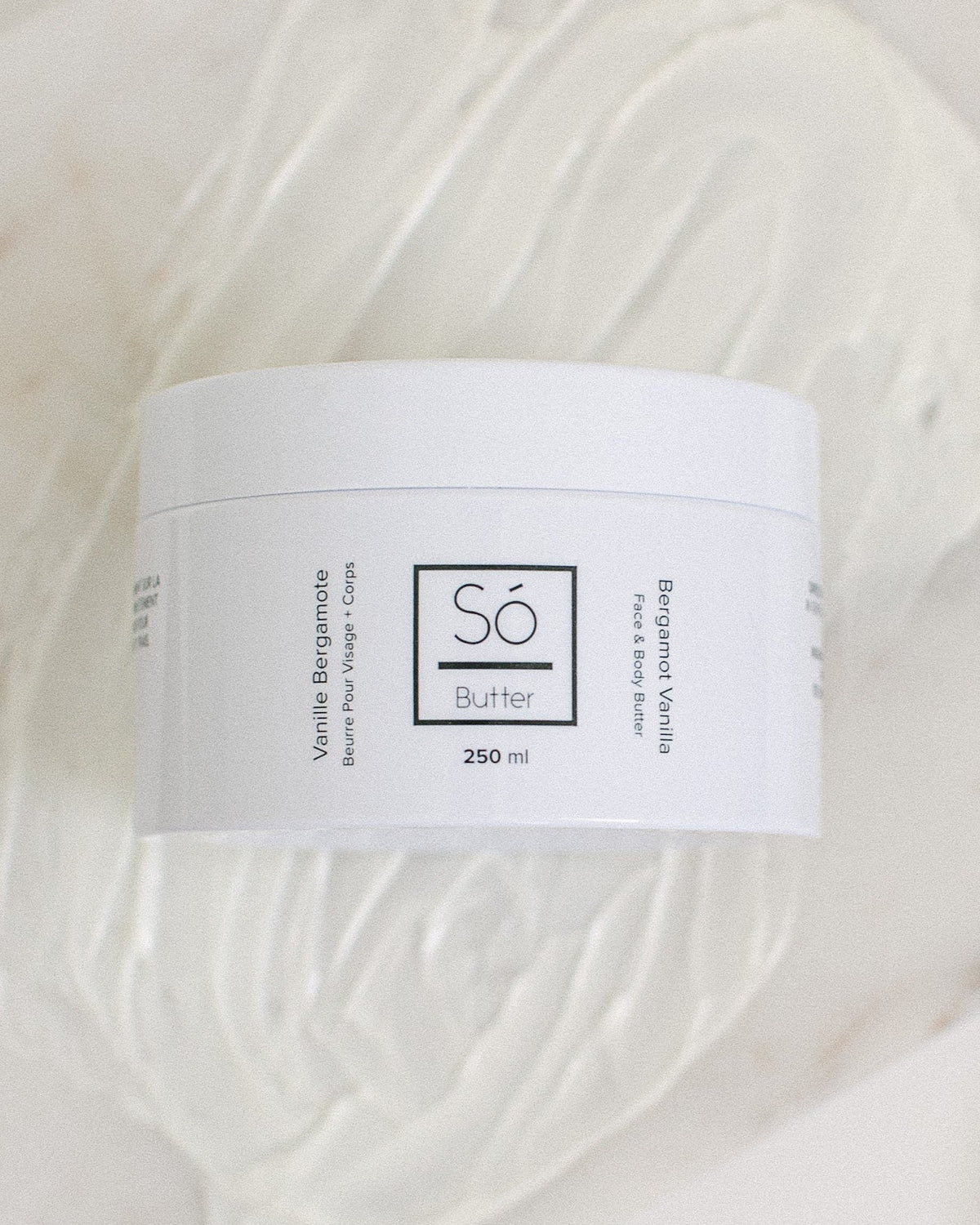 A white plastic jar of Só Butter Bergamot Vanilla body butter, placed on top of a textured background of white whipped product