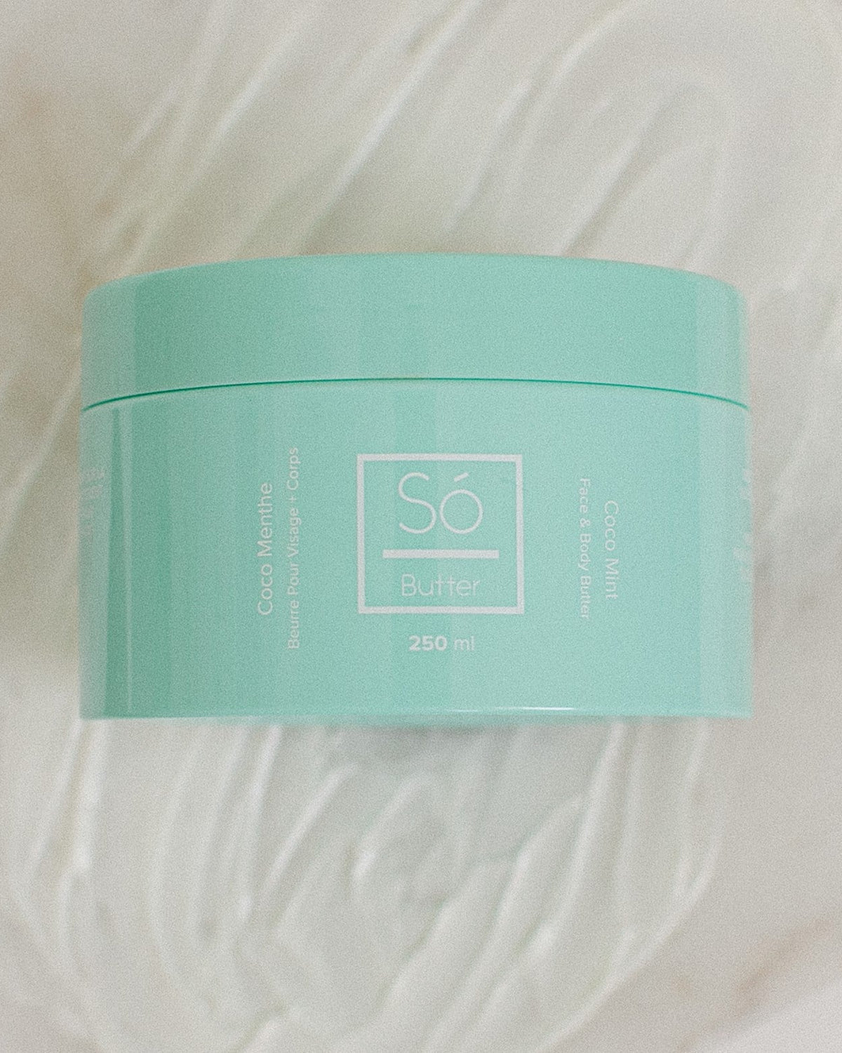 A mint green plastic jar of Só Butter Coco Mint body butter, placed on top of a textured background of white whipped product