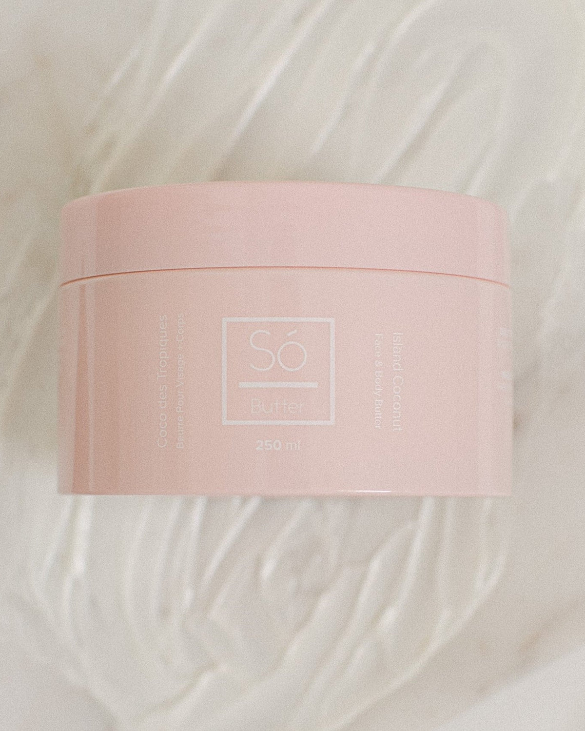 A pink plastic jar of Só Butter Island Coconut body butter, placed on top of a textured background of white whipped product