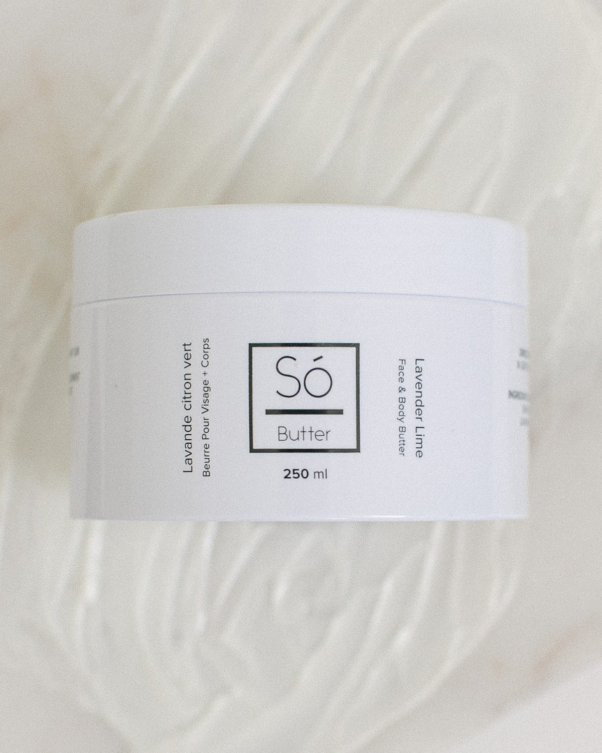 A white plastic jar of Só Butter Lavender Lime body butter, placed on top of a textured background of white whipped product