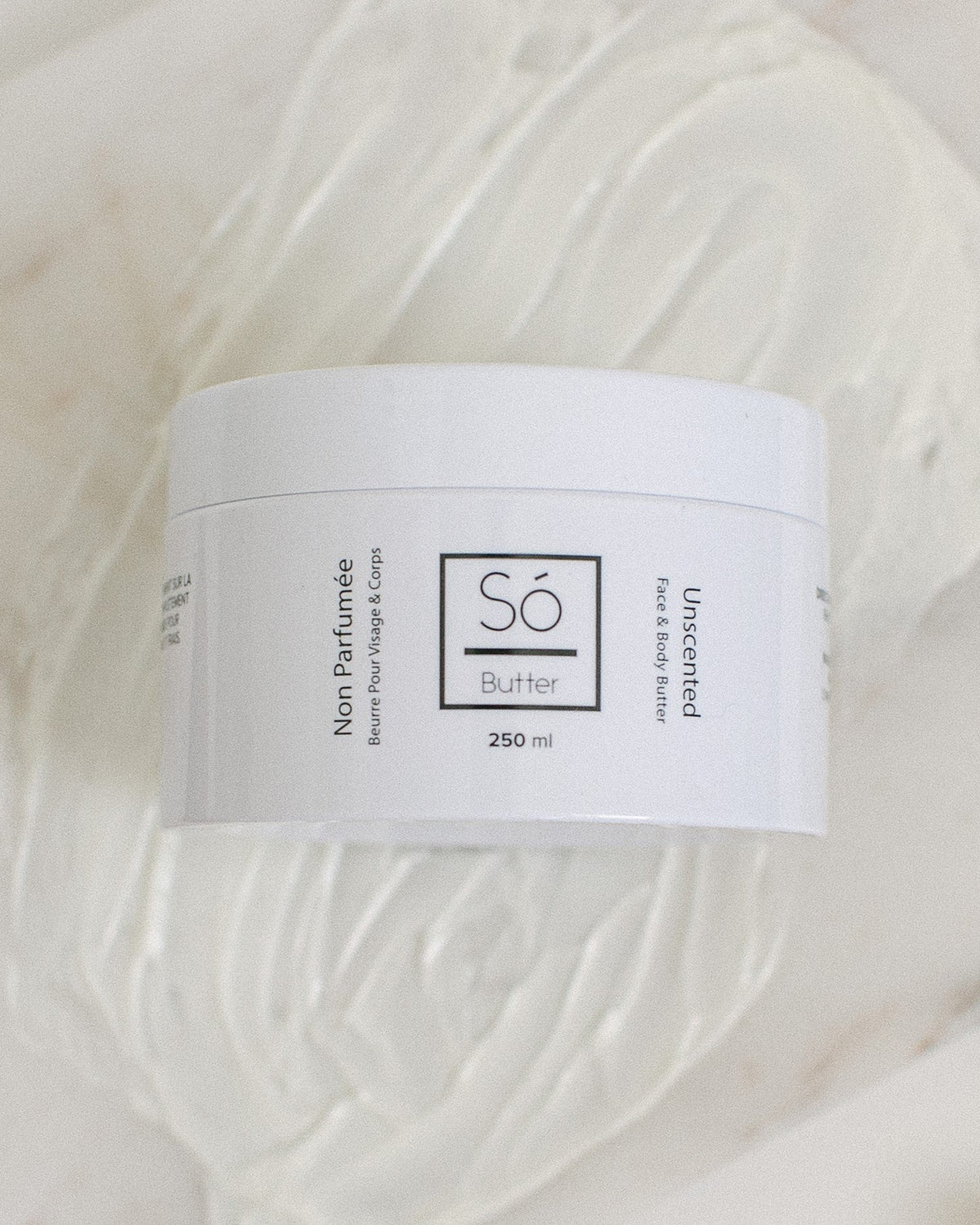 A white plastic jar of Só Butter Unscented  body butter, placed on top of a textured background of white whipped product