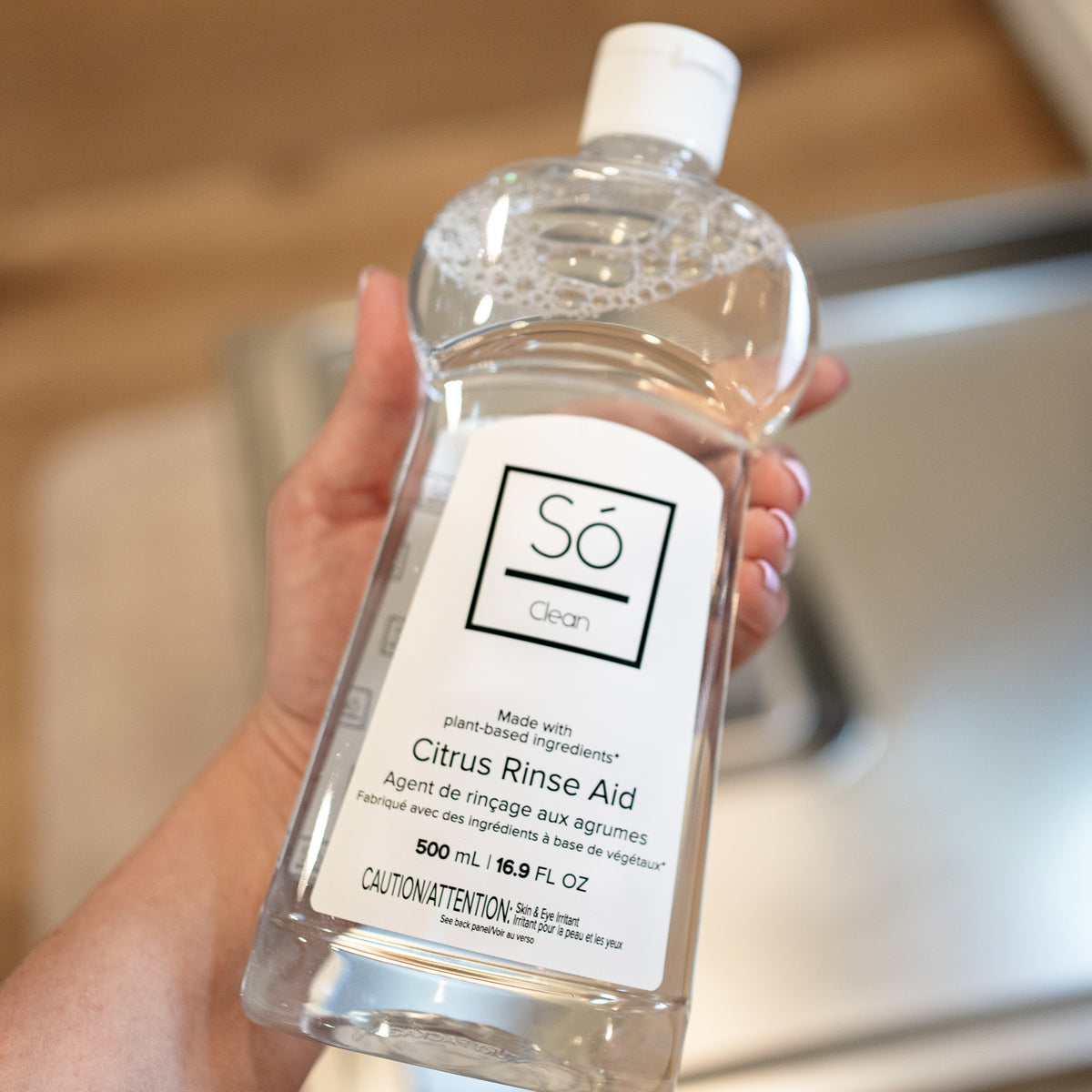 A hand holding a clear bottle with a clear liquid and white Só branded label listing the product as Citrus Rinse Aid, an open dishwasher can be seen in the blurred background.