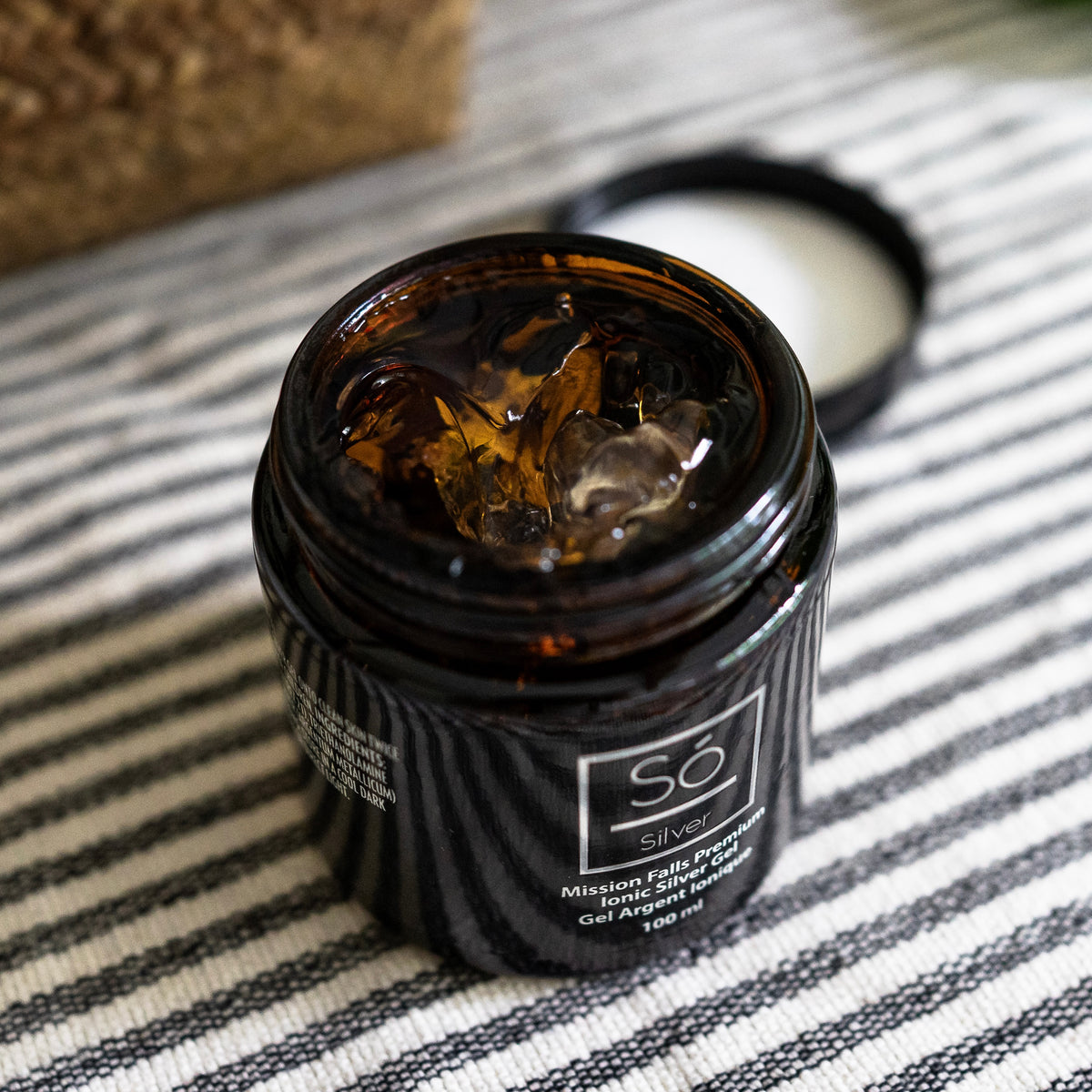 An open jar of Só Silver Mission Falls Ionic Silver Gel 100 ml on a black and white striped fabric surface, with the lid resting beside it.