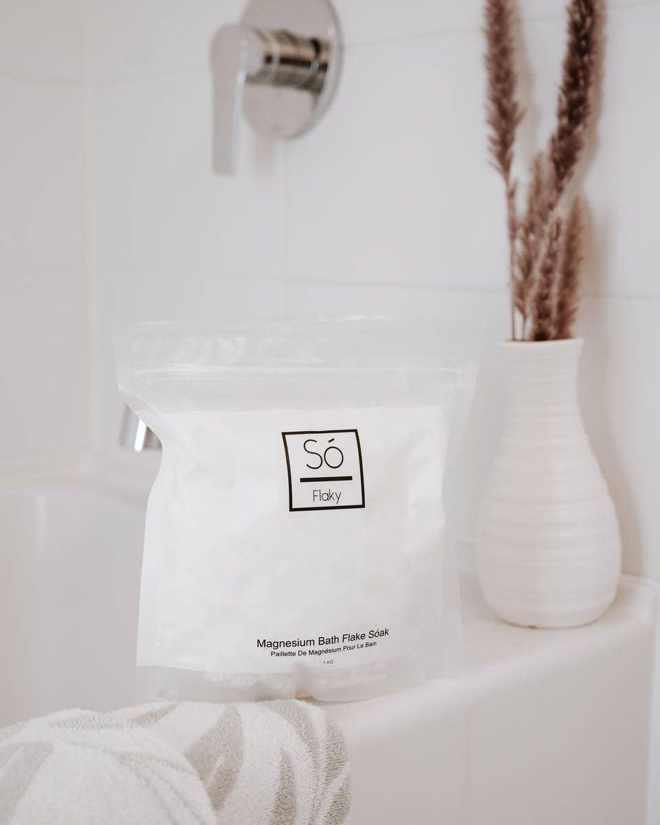 White bag with 'Só Flaky' branding on the edge of a bathtub with a cream coloured vase with dried florals.