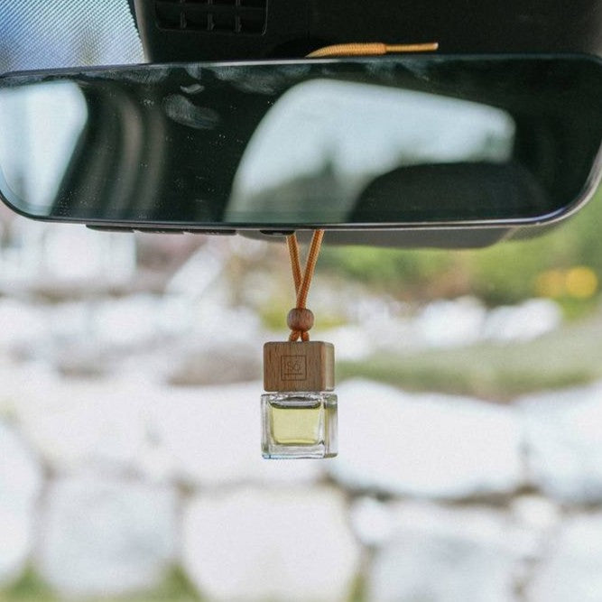 Small square glass jar with a Só branded wooden lid and wood bead with tan coloured shoelace-like string shown hanging from a car rearview mirror filled with a light yellow liquid