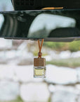 Small square glass jar with a Só branded wooden lid and wood bead with tan coloured shoelace-like string shown hanging from a car rearview mirror filled with a light yellow liquid