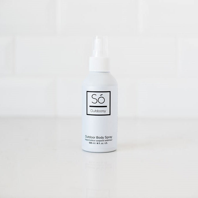 A white bottle of Só Outdoorsy Outdoor Body Spray with a white nozzle cap, standing on a white surface against a white tiled background.