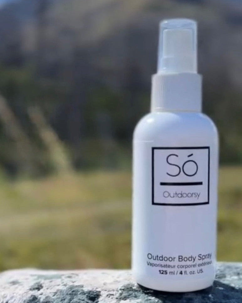 A white bottle of Só Outdoorsy outdoor body spray standing on a gray stone surface with a blurred mountain landscape in the background.