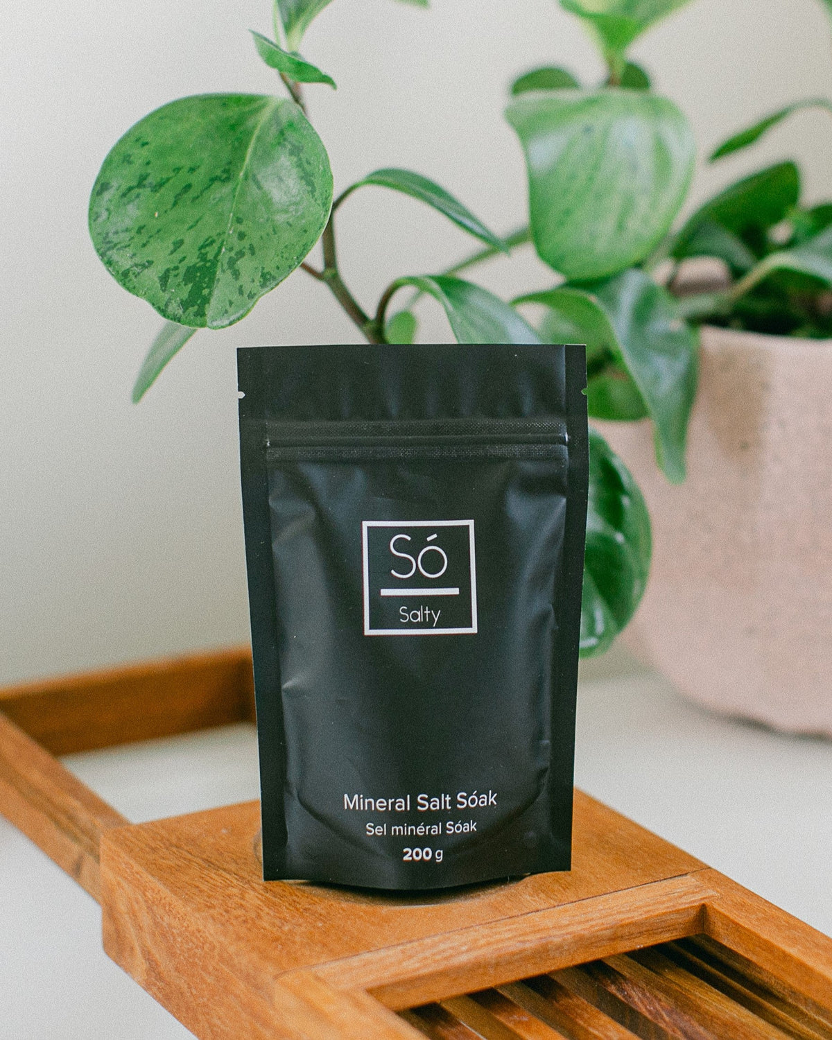 A black package of Só Salty (Essential Oil) bath soak with Epsom salt and Himalayan Pink salt, scented with various essential oils, displayed on a wooden bath tub tray with a plant in a pink pot behind it.
