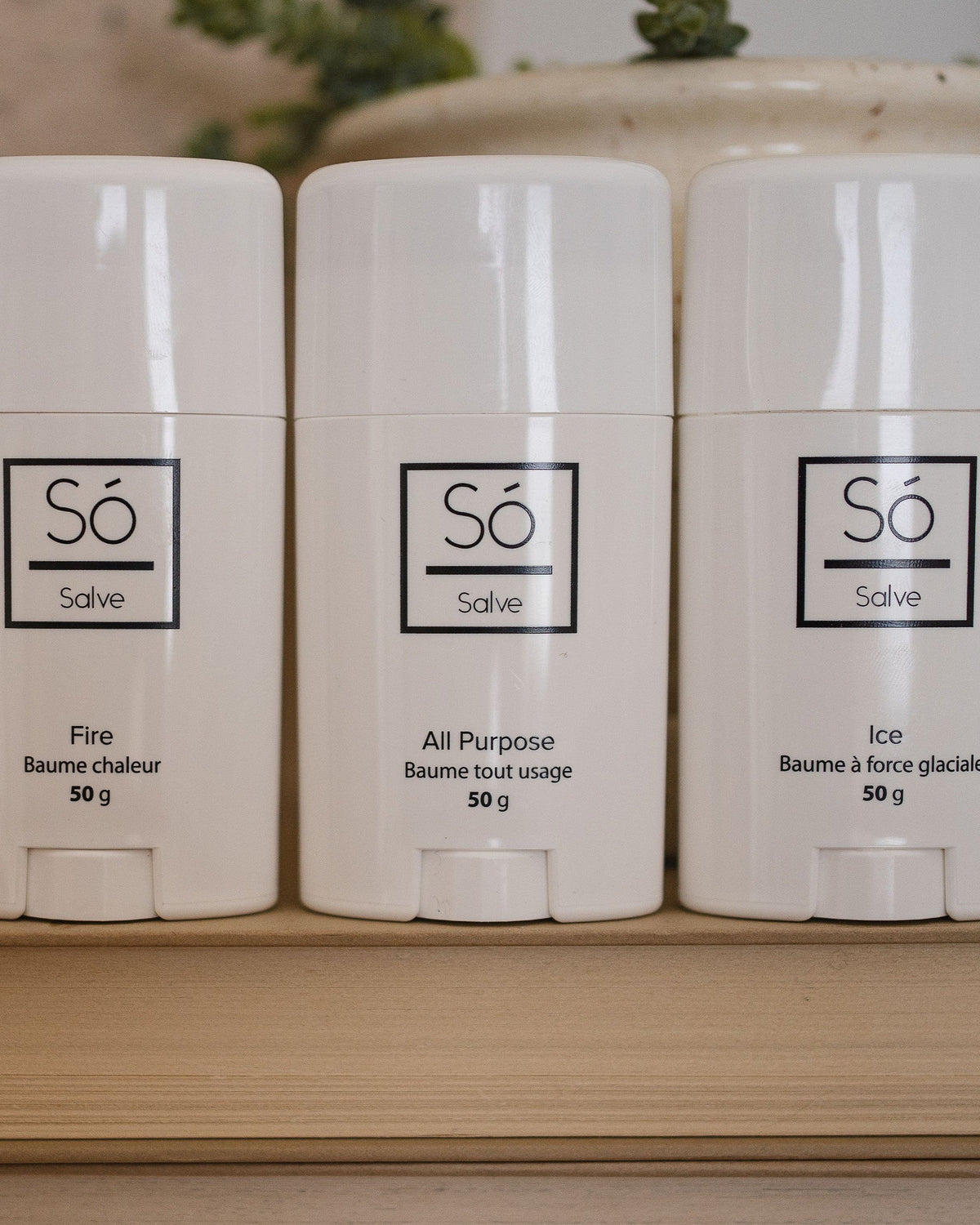Só Luxury Collection of Salves featuring Fire warming salve Ice cooling salve and All purpose healing salve in 50 g twist-up sticks standing on a book with greenery in the background.
