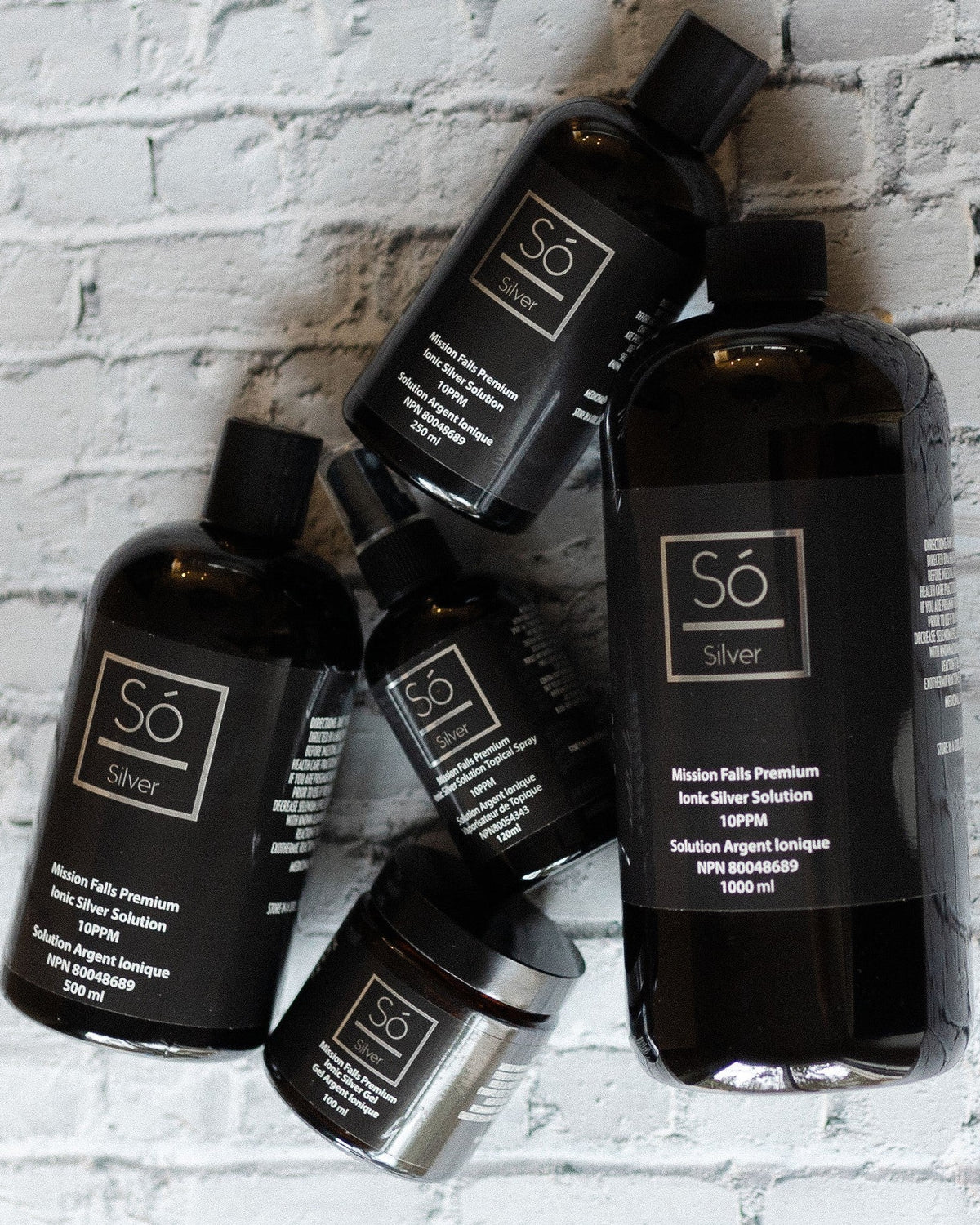 Four black bottles of various size and a black glass jar all labeled Só Silver Mission Falls Ionic Silver Solution styled on a white brick background.