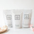 Three white stand-up pouches of Só skincare products arranged on a white surface: Coco Cream Soak, Blush Soak, and Coconut & Oat Mix Bath. A wooden spoon with bath powder sits at left, and pale pink flowers are at right, against a clean white background.