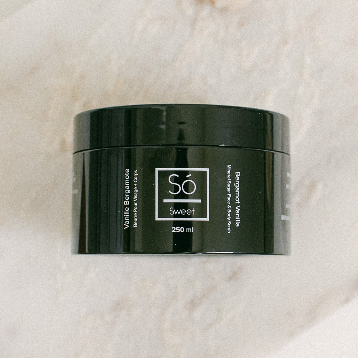 A 250 ml black jar of Só Sweet Bergamot Vanilla body scrub, resting on a light beige marble surface.