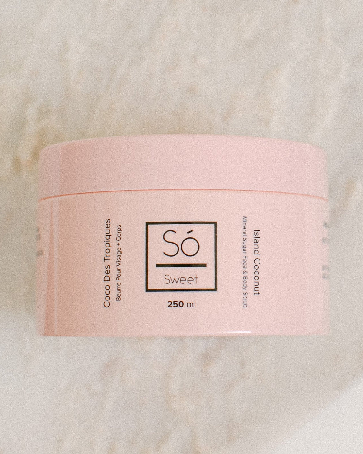 A 250 ml pink jar of Só Sweet Island Coconut body scrub, resting on a light beige marble surface.