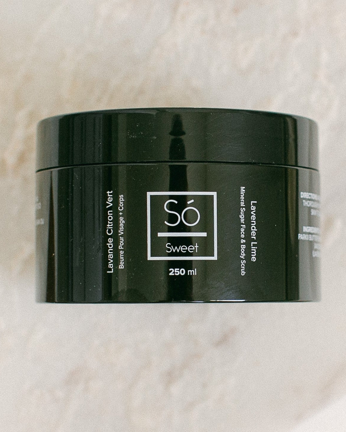 A 250 ml black jar of Só Sweet Lavender Lime body scrub, resting on a light beige marble surface.