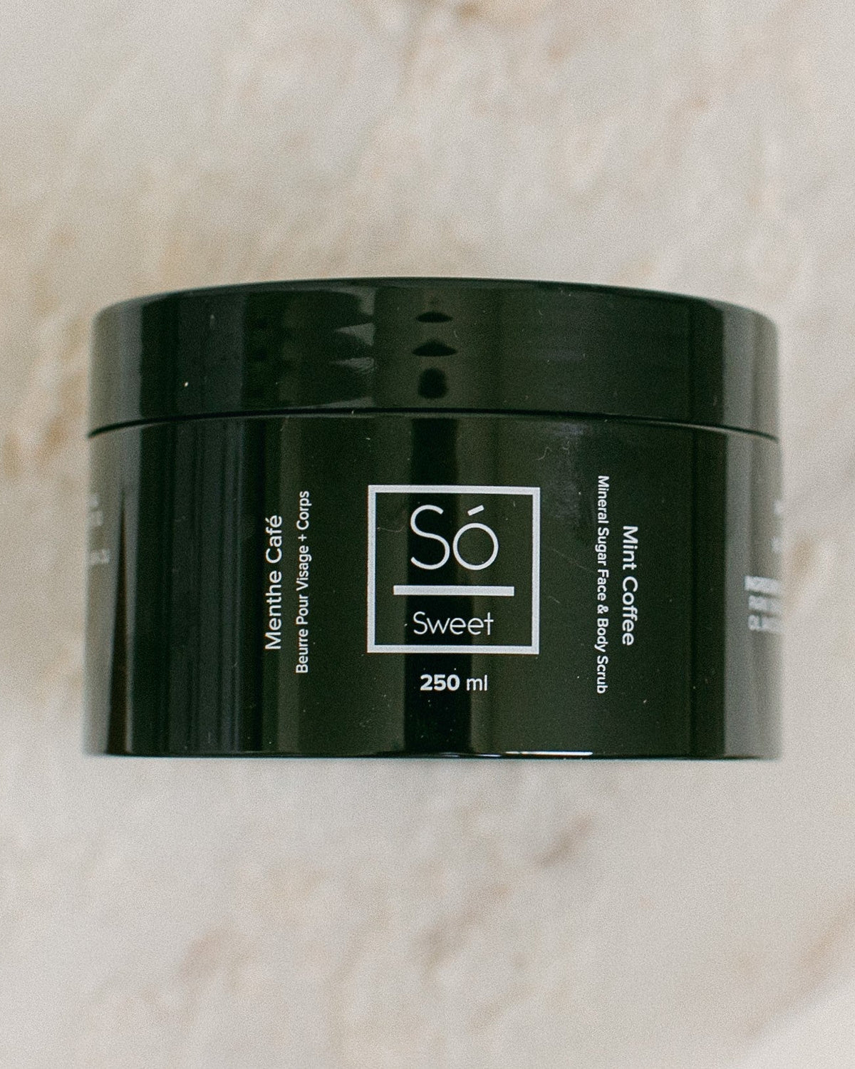 A 250 ml black jar of Só Sweet Mint Coffee body scrub, resting on a light beige marble surface.