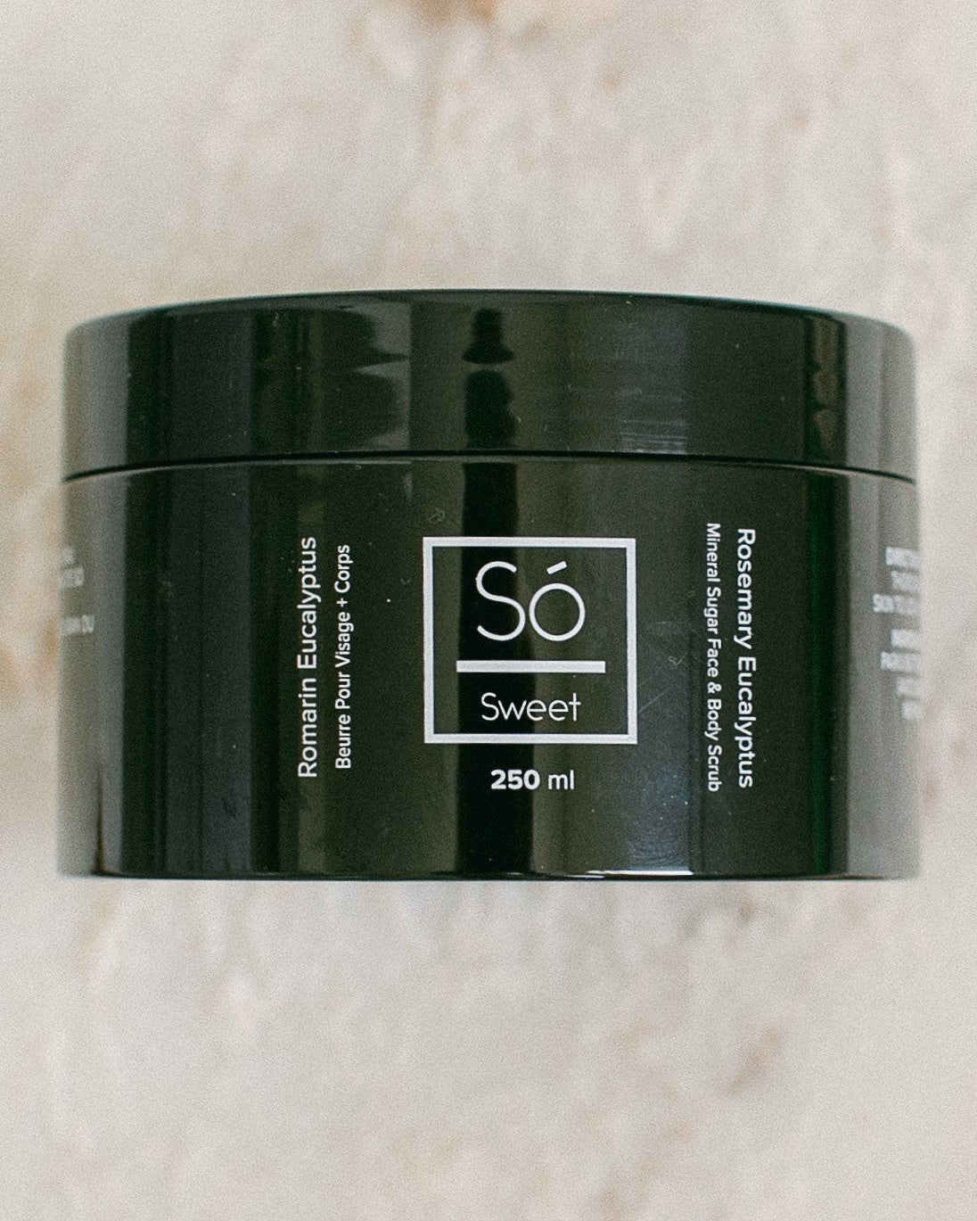 A 250 ml black jar of Só Sweet Rosemary Eucalyptus body scrub, resting on a light beige marble surface.