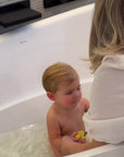 A mom in a white blouse with blonde hair bathing a blonde baby in a white bathtub using a konjac sponge to wash