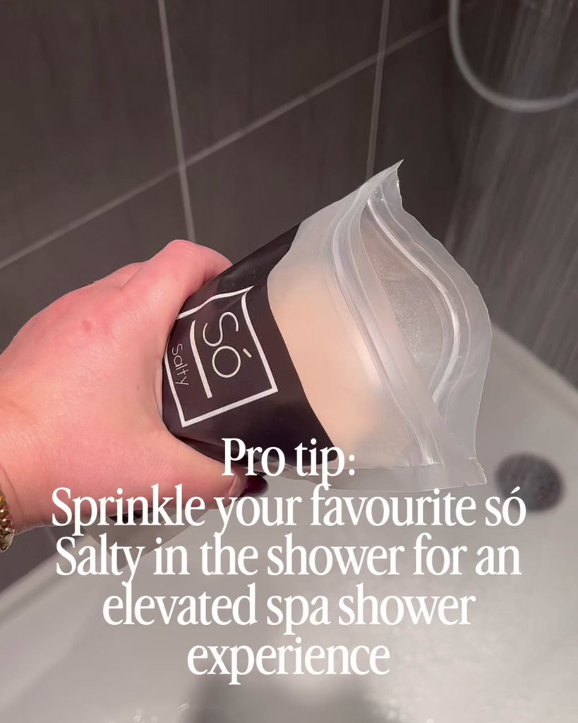 A video depicting a hand holding a bag of Só Salty in a shower with the water running and sprinkling some pink salt cystals onto the shower floor. Text on screen says "Pro tip: Sprinkle your favourite só salty in the shower for an elevated spa shower experience"