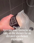 A video depicting a hand holding a bag of Só Salty in a shower with the water running and sprinkling some pink salt cystals onto the shower floor. Text on screen says "Pro tip: Sprinkle your favourite só salty in the shower for an elevated spa shower experience"