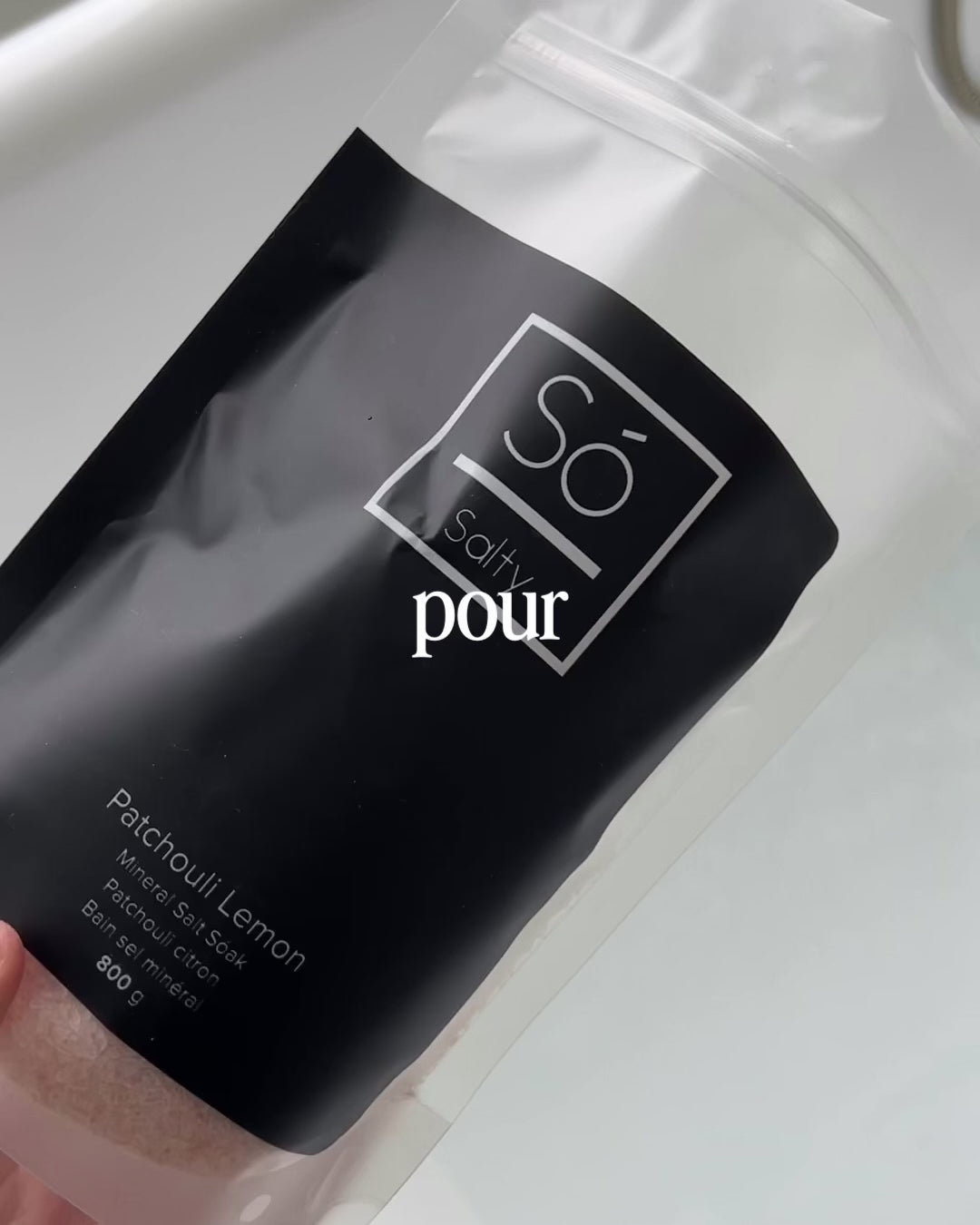A video depicting a hand holding an open bag of Só Salty Patchouli Lemon over a filling bathtub and pouring pink salt into the tub. Text overlay says "pour. sóak. unwind"