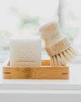 A bamboo soap dish holding a white bar of soap embossed with the brand 'Só' and a small wooden-handled cleaning brush with natural bristles, set on a bright white surface near a softly blurred window.