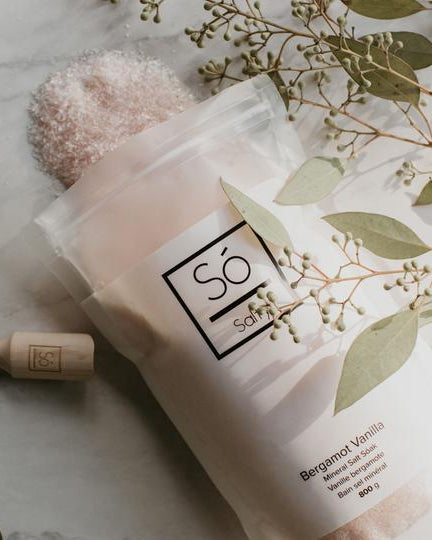 A package of Só Salty bath soak with Epsom salt and Himalayan pink salt labelled Bergamot Vanilla styled with a wooden scoop, pink salt cystals spilling out of the bag and eucalyptus branches.