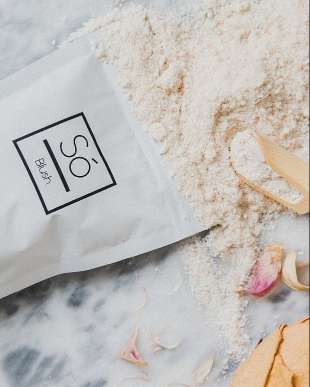 A package of Só Luxury brand Blush soak with a light pink powdered product scattered around, presented on a marble surface with rose petals.