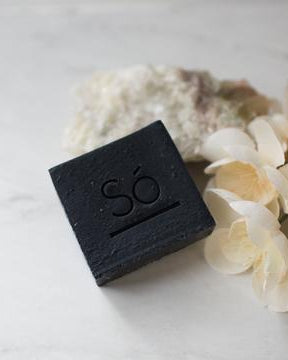 A black bar of soap branded with Só styled on a marble background with natural stone and faux florals.