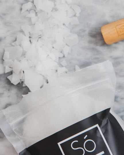 A bag of Só brand magnesium bath flakes spilled open with the contents, a white crystalline substance, scattered on a grey marble surface with a wooden Só branded scoop.