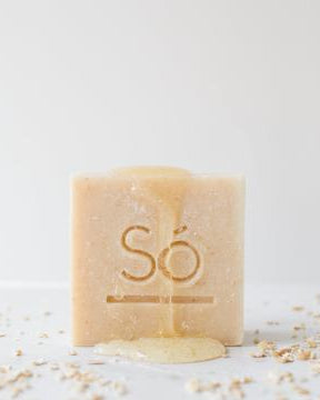 A beige bar of soap branded Só standing upright with honey dripping down the front and oats sprinkled around on a white background.