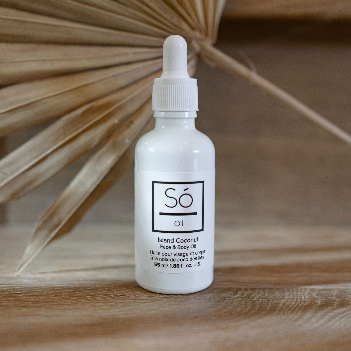 A white bottle with a dropper cap labeled 'Só Oil' with a scent described as Island Coconut. It is placed on a wooden surface with a dried palm leaf in the background.