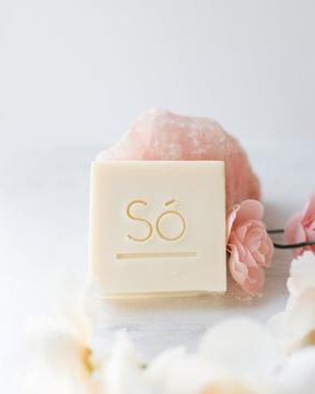 A cream coloured square bar of soap branded with Só styled on a light coloured background with a rose quartz stone and faux florals.
