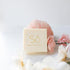 A cream coloured square bar of soap branded with Só styled on a light coloured background with a rose quartz stone and faux florals.