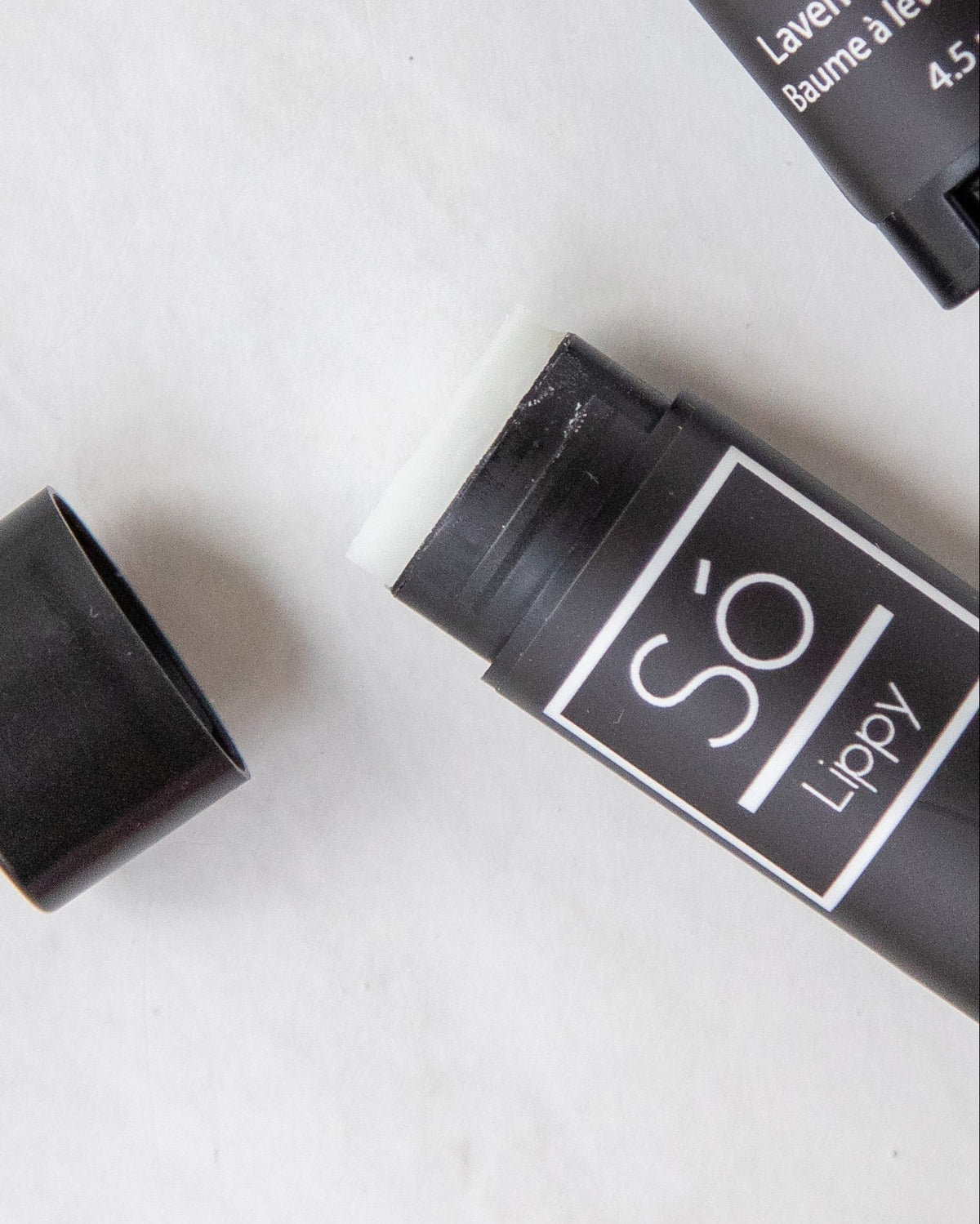 A stick of lip balm with the brand name Só Lippy placed on a surface, open, with its cap next to it, against a marble background.