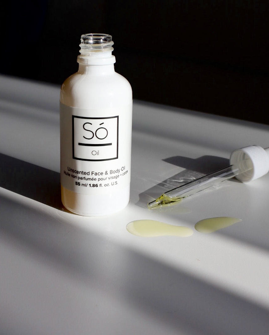 A white bottle labelled Só Oil Unscented Face & Body Oil with the dropper  laying beside it containing oil, placed on a surface with a few drops of oil visible in front of the bottle.