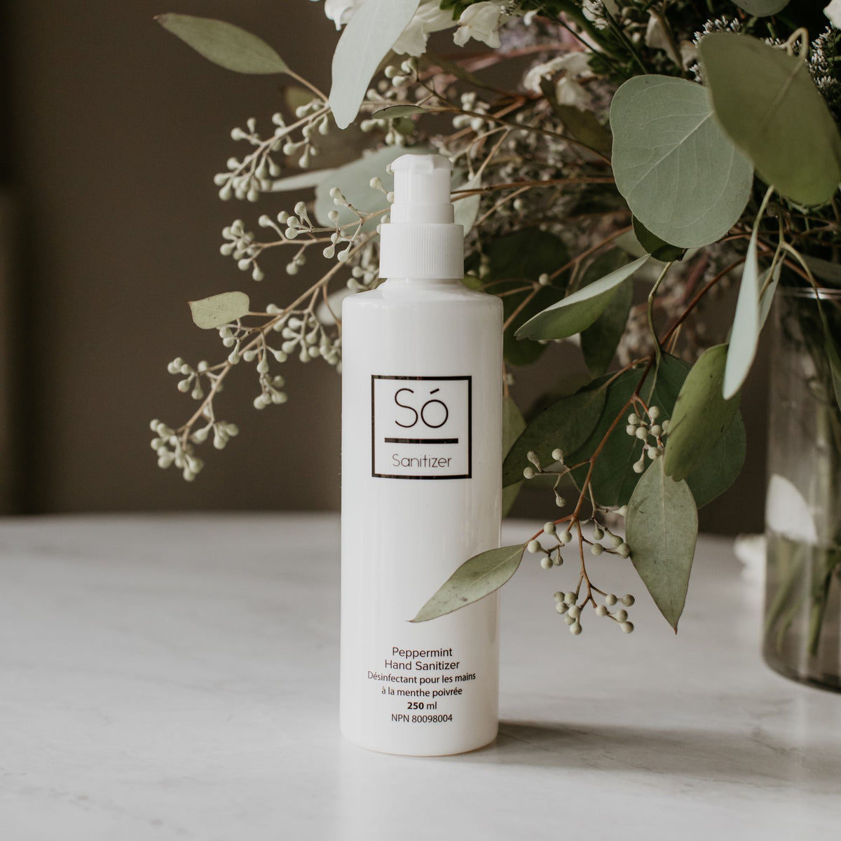 A white bottle of branded 'Só Sanitizer' with a pump top styled on a marble surface with natural greenry