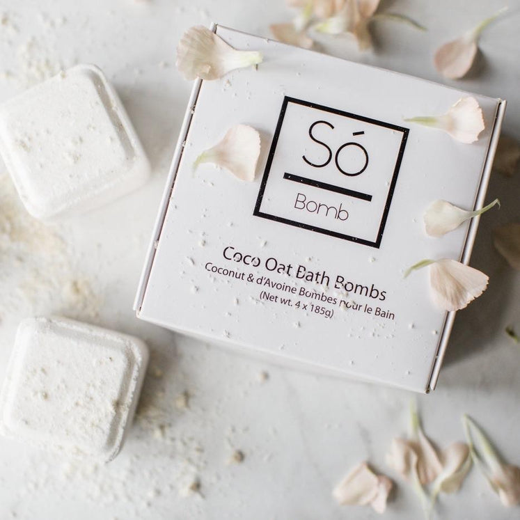 A bath bomb from Só Bomb with packaging that reads Coco Oat Bath Bombs' The product is displayed on a white marble backdrop with light pink flower petals and completed bath bombs with a powdered substance sprinkled on both the completed bath bombs and the branded box.