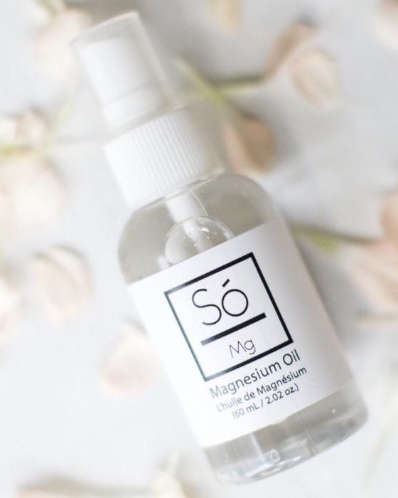 A clear spray bottle of Só Magnesium Oil with a white pump and white label, photographed on a light surface with soft blurred flowers around it.