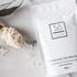 A package of Só Luxury Coco Oat Milk Bath powder with some scattered on a gold spoon beside it, all placed on a white marble surface.