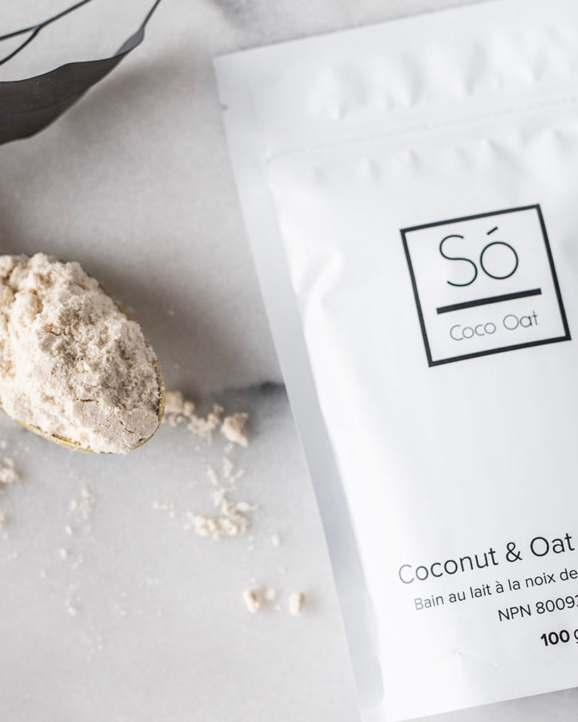 A package of Só Luxury Coco Oat Milk Bath powder with some scattered on a gold spoon beside it, all placed on a white marble surface.