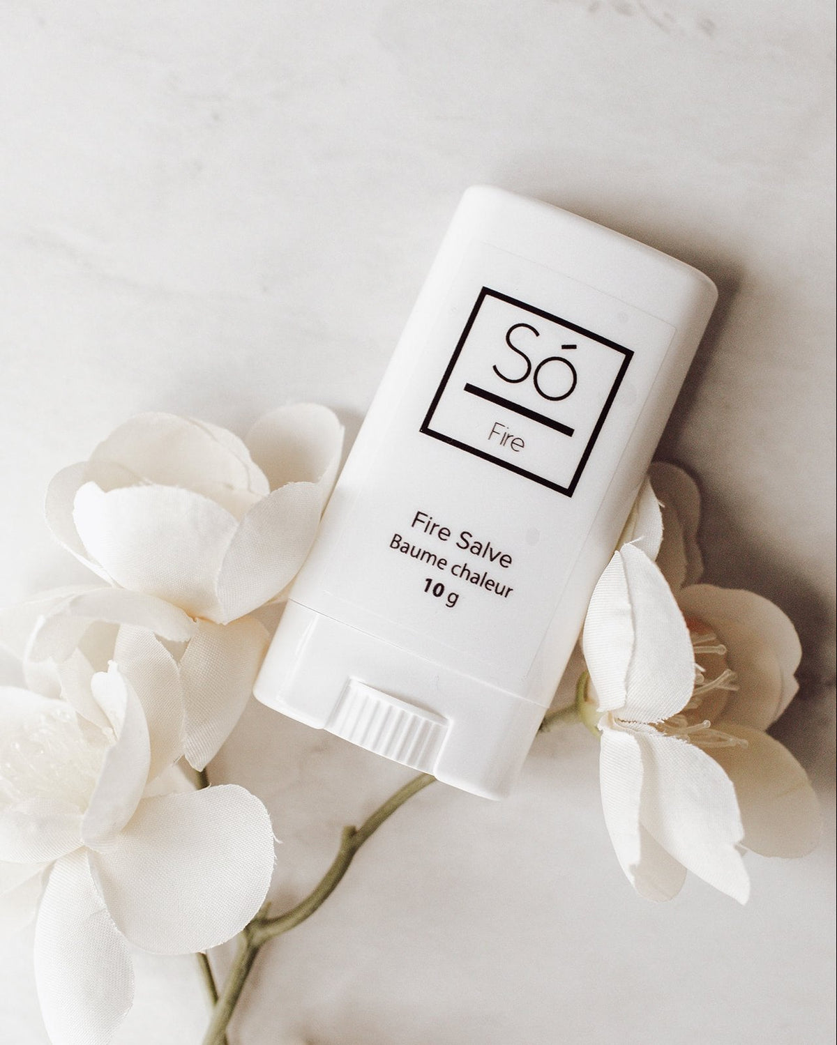 A white plastic deodorant style container branded Só Salve Fire Salve a warming muscle salve in 10 g twist-up stick styled on a marble background with white flowers.