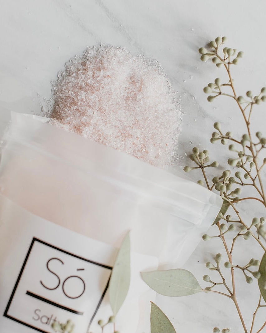 A frosted resealable pouch labeled Só salty with pale pink salt crystals spilling out, styled on a light marble surface with eucalyptus branches.