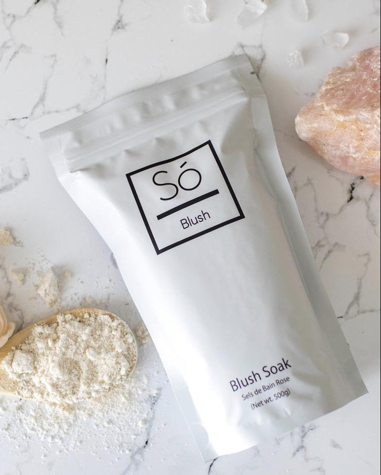A white stand-up pouch of Só Blush Blush Soak set on a white marble surface with scattered powder, a spoonful of the powder and a decorative pink stone.