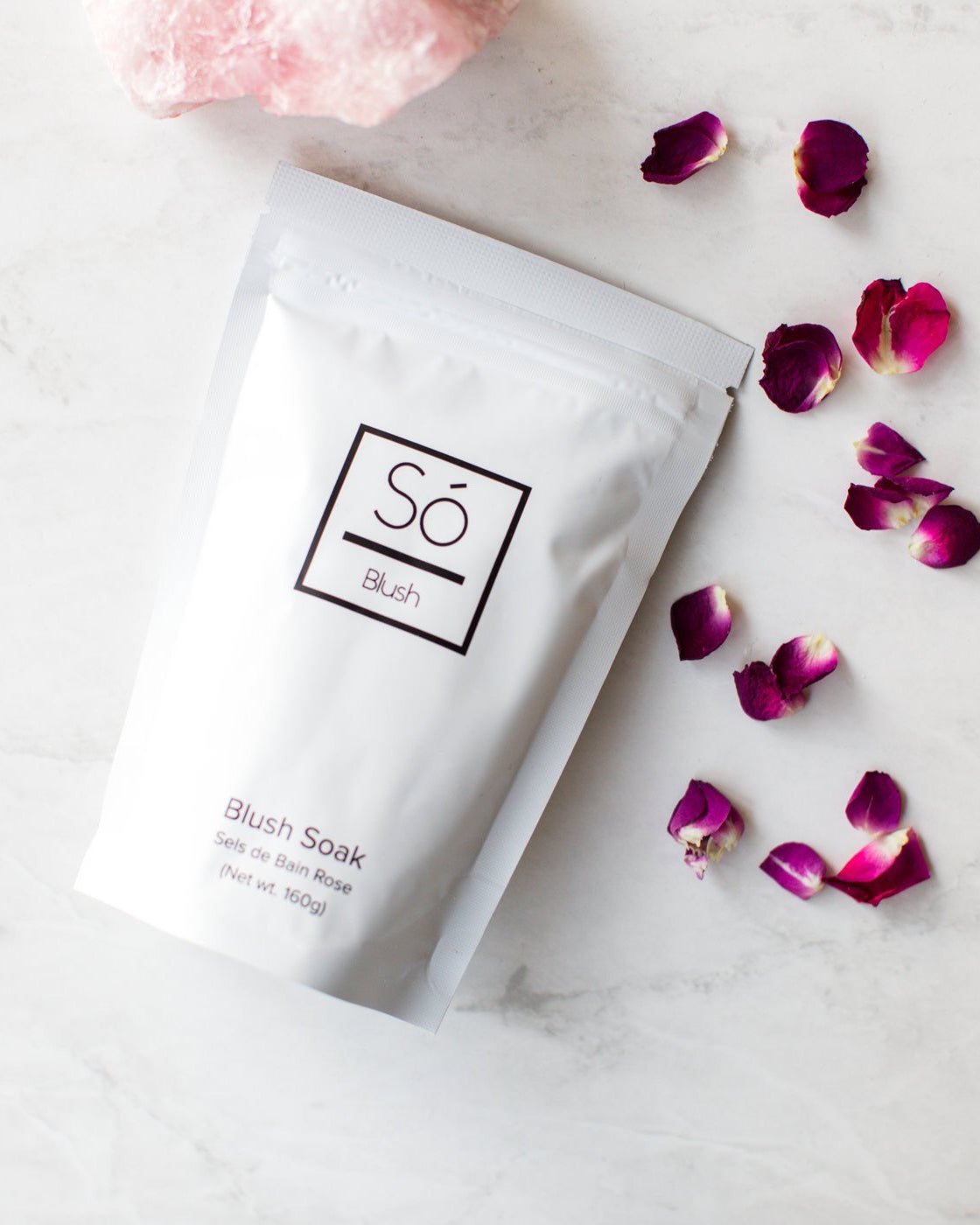 A package of Blush Sóak bath soak by Só Luxury, with a decorative pink stone and rose petals scattered around on a marble surface.