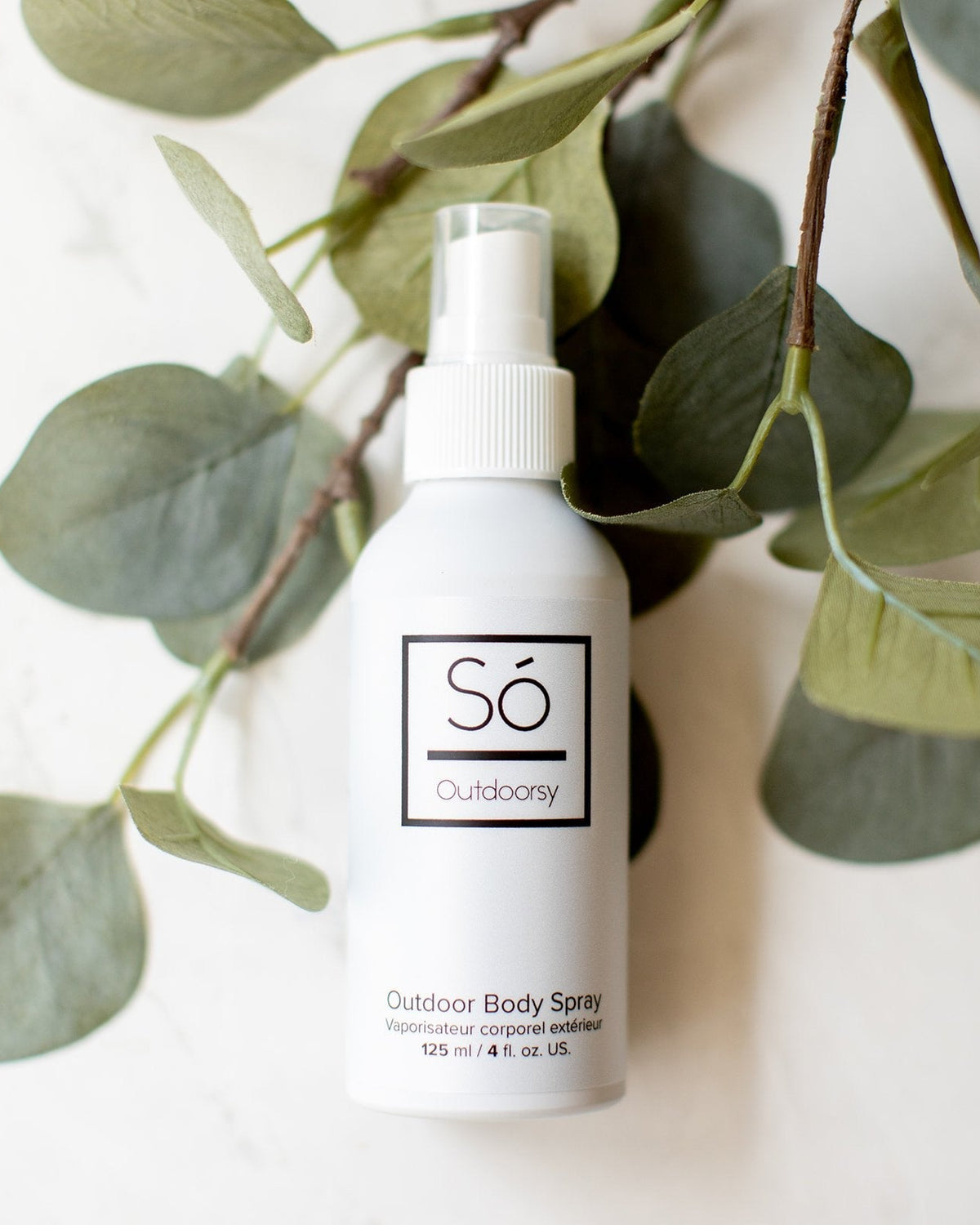 A white spray bottle labeled 'Só Outdoorsy' placed against a backdrop of green leaves, representing an outdoor body spray product.