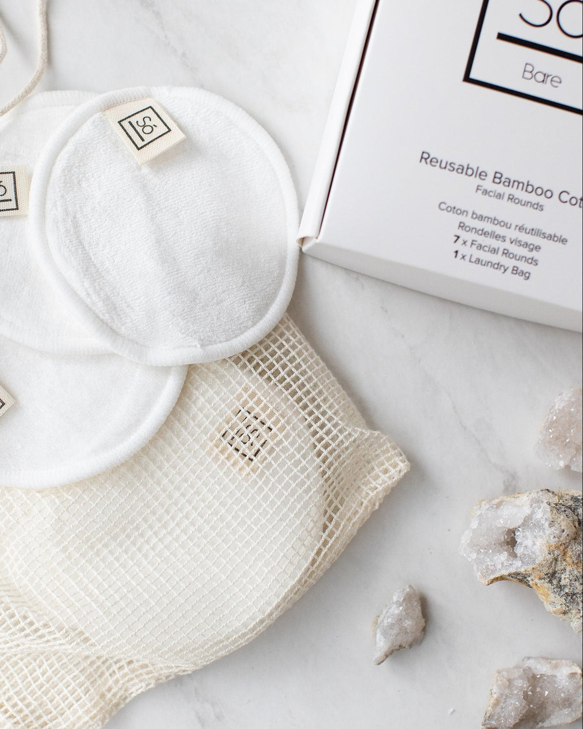 White reusable cotton face pads with small sewn-on 'Só' branded tags, arranged on a cream mesh bag next to a white box on a light marble surface with a few crystals.
